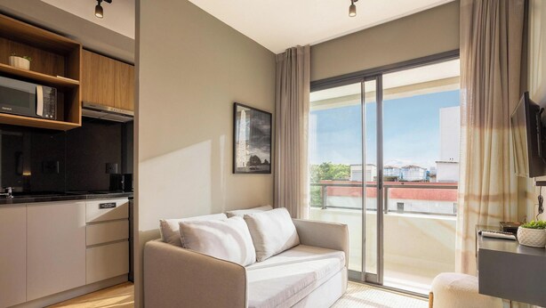 A modern living room with a sofa, a flat-screen TV, a kitchen area with a microwave, and a large sliding glass door leading to an outdoor balcony. - Condo. w/ Rooftop Pool, 270m fr/ the Metro, Jardins - Gabs (São Paulo)