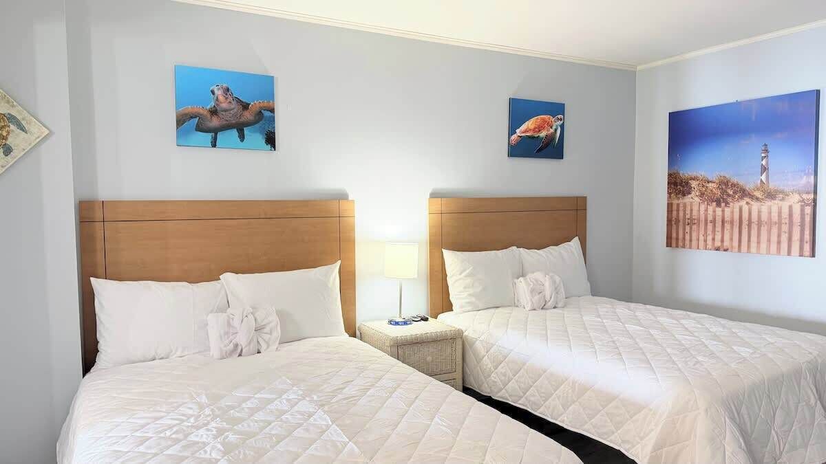 A hotel room with two beds, a nightstand, and wall art depicting animals and a lighthouse.