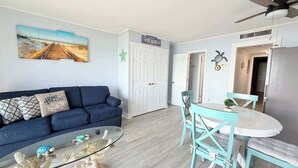 Ein Wohnzimmer mit einem blauen Sofa, einem Glastisch und einem Essbereich mit türkisfarbenen Stühlen und einem weißen Tisch.