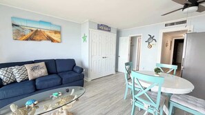 Ein Wohnzimmer mit einem blauen Sofa, einem Glastisch und einem Essbereich mit türkisfarbenen Stühlen und einem weißen Tisch.
