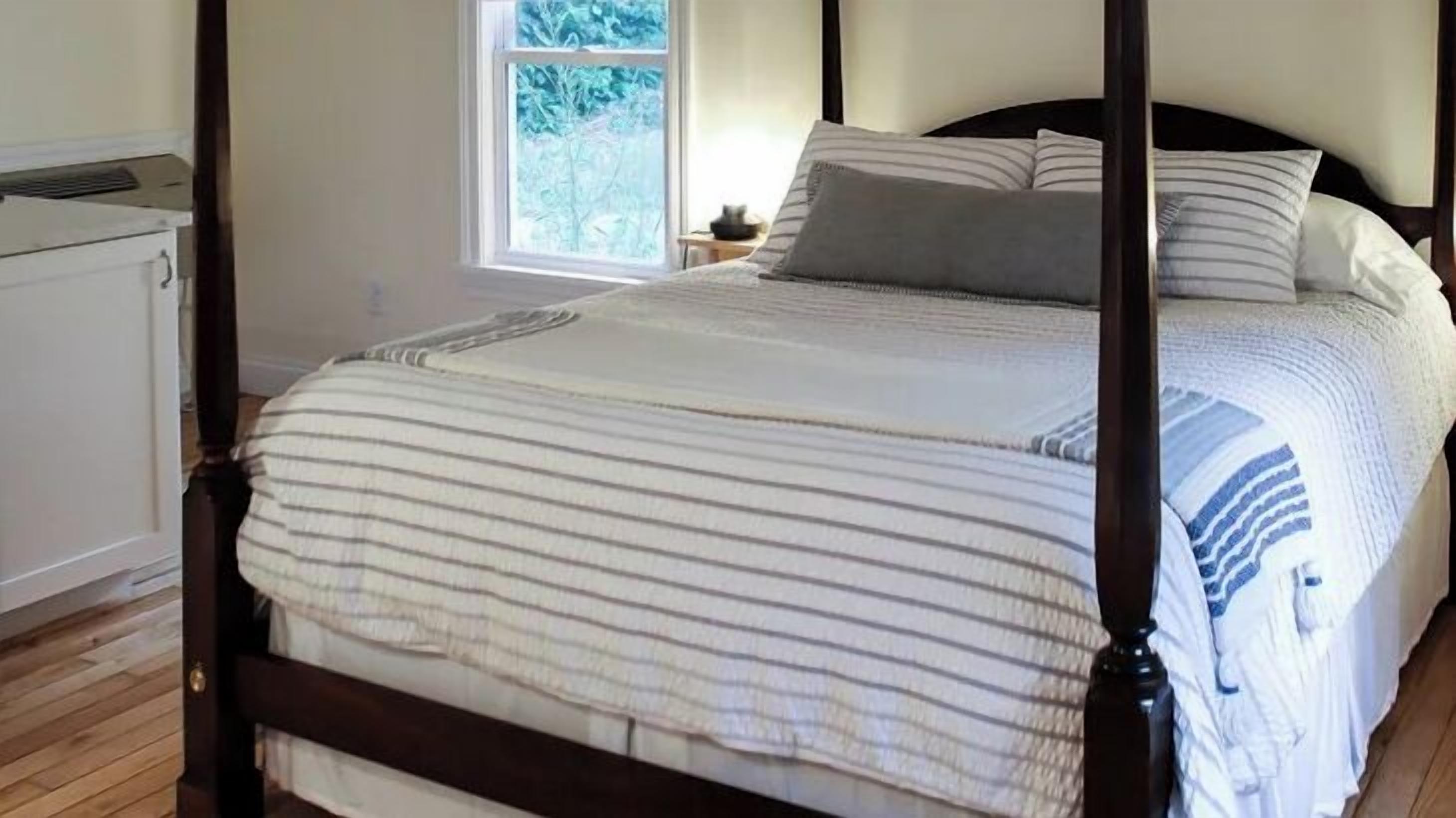 A four-poster bed with a striped bedspread, a window with a view of greenery, and a bedside table with a lamp.