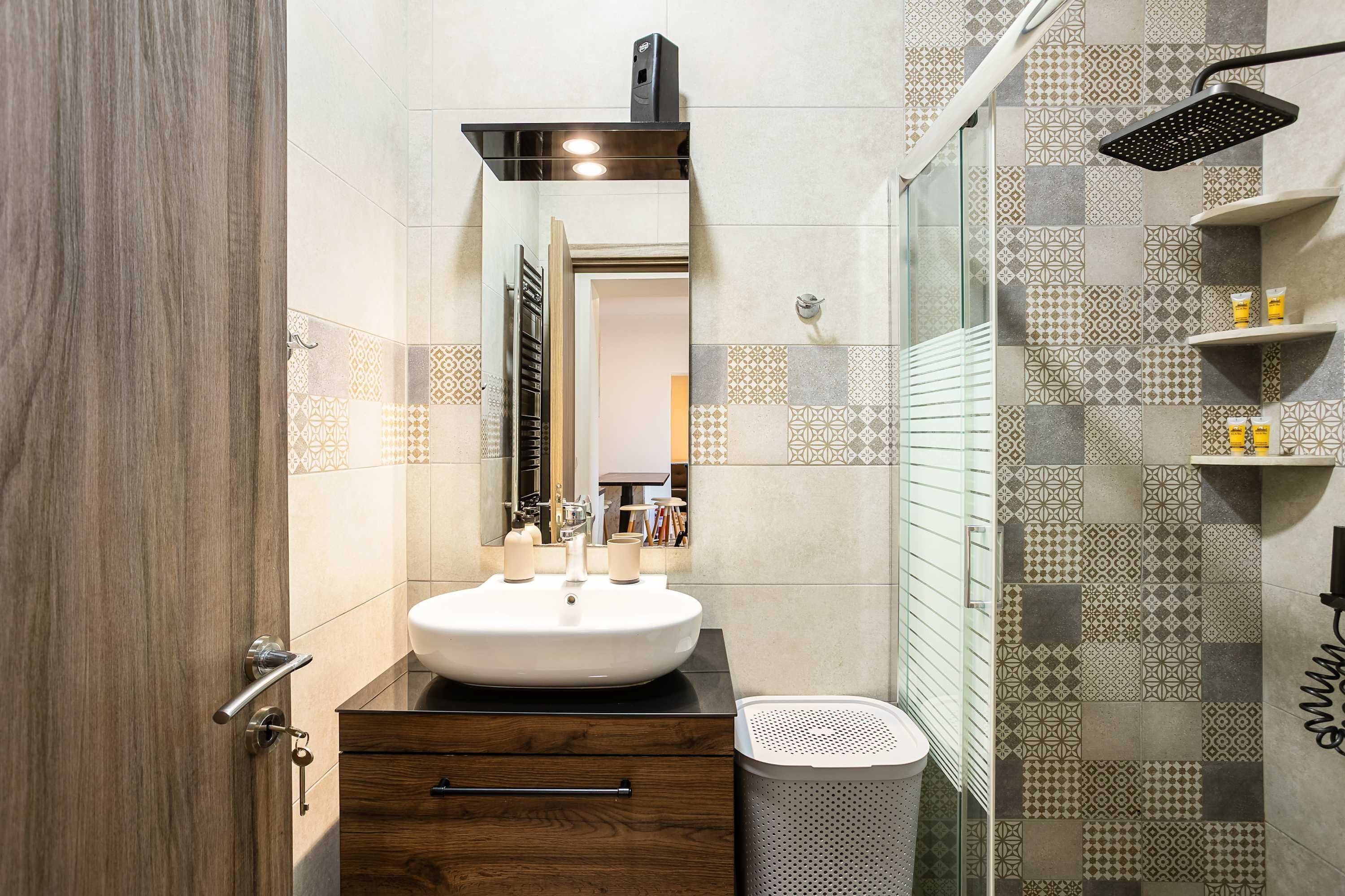 A modern bathroom with a wooden vanity, a white sink, a mirror, and a shower with a patterned tile wall.