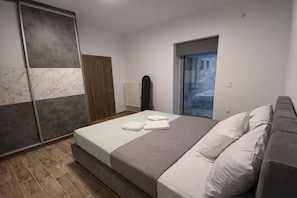Quarto com cama, guarda-roupa, radiador e vista para o céu azul e um edifício.