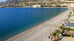 A coastal beach with a pebble beach, a promenade lined with palm trees, and buildings in the background. - AG Stone Home - Idyllic Beachside Villa in Isthmia (Loutraki-Agioi Theodoroi)