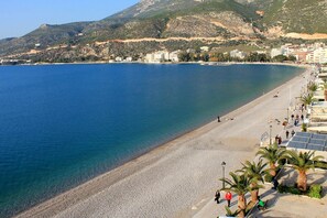 A coastal beach with a pebble beach, a promenade lined with palm trees, and buildings in the background. - AG Stone Home - Idyllic Beachside Villa in Isthmia (Loutraki-Agioi Theodoroi)
