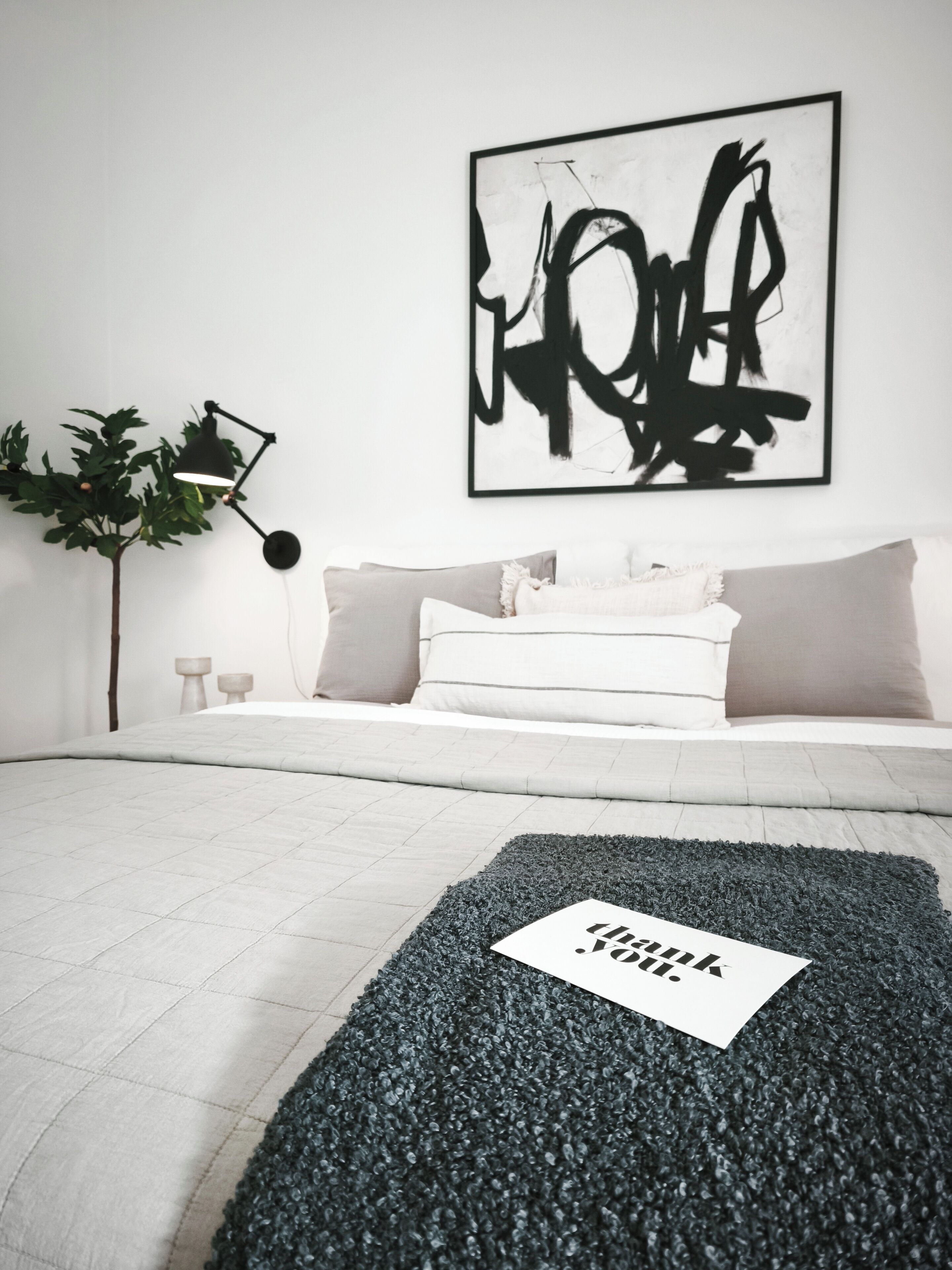 A bedroom with a bed, a black abstract painting, a plant, and a desk lamp.
