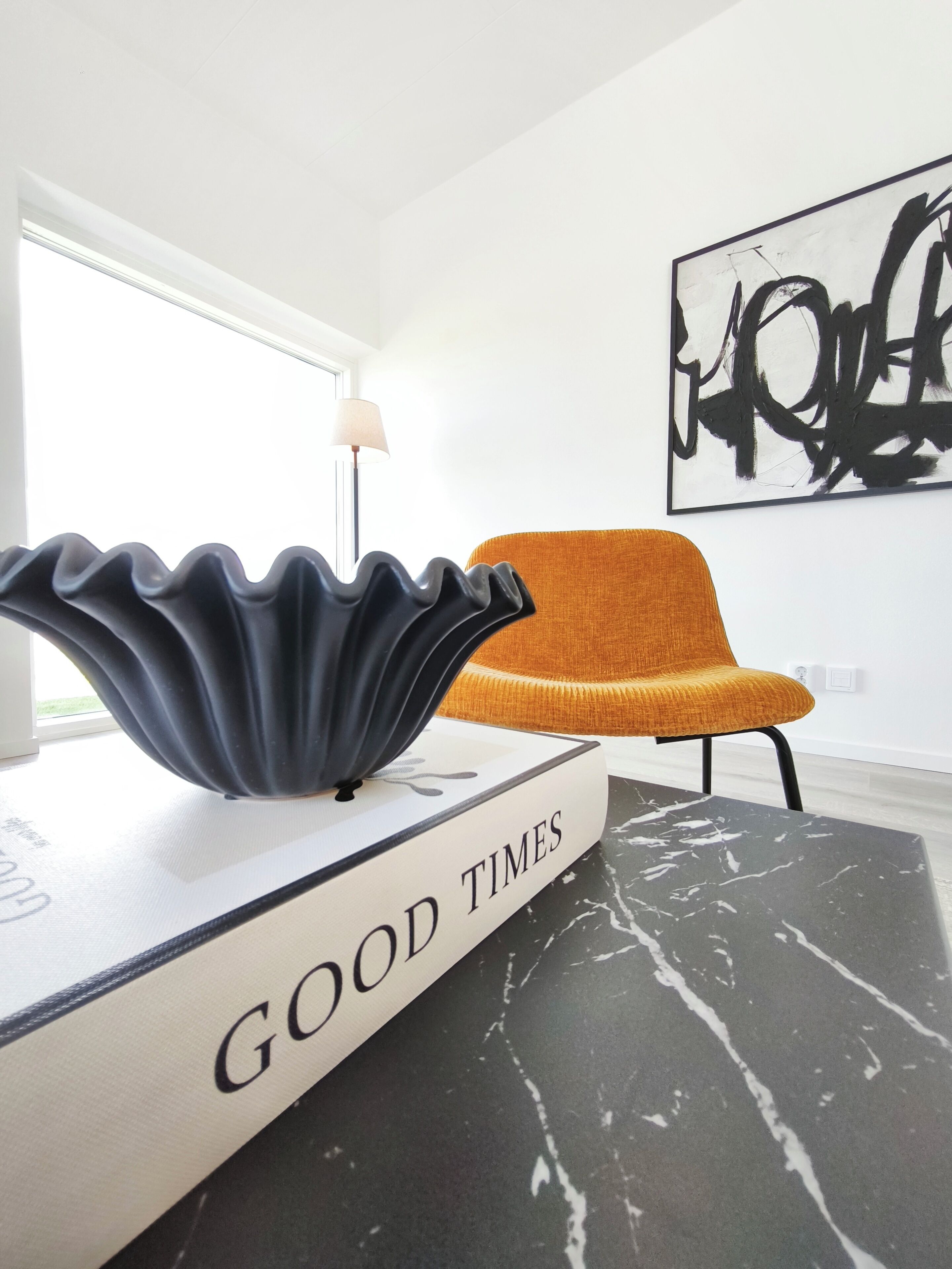 Un interior moderno con una mesa de mármol blanco y negro, un libro titulado 'GOOD TIMES' encima, un cuenco decorativo negro, una silla naranja y un cuadro abstracto blanco y negro en la pared.
