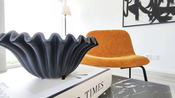 Un interior moderno con una mesa de mármol blanco y negro, un libro titulado 'GOOD TIMES' encima, un cuenco decorativo negro, una silla naranja y un cuadro abstracto blanco y negro en la pared.