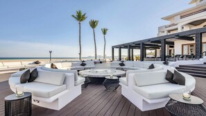 Eine Dachterrasse mit weißen Sofas und einem zentralen Couchtisch, mit Blick auf einen Strand mit Palmen.