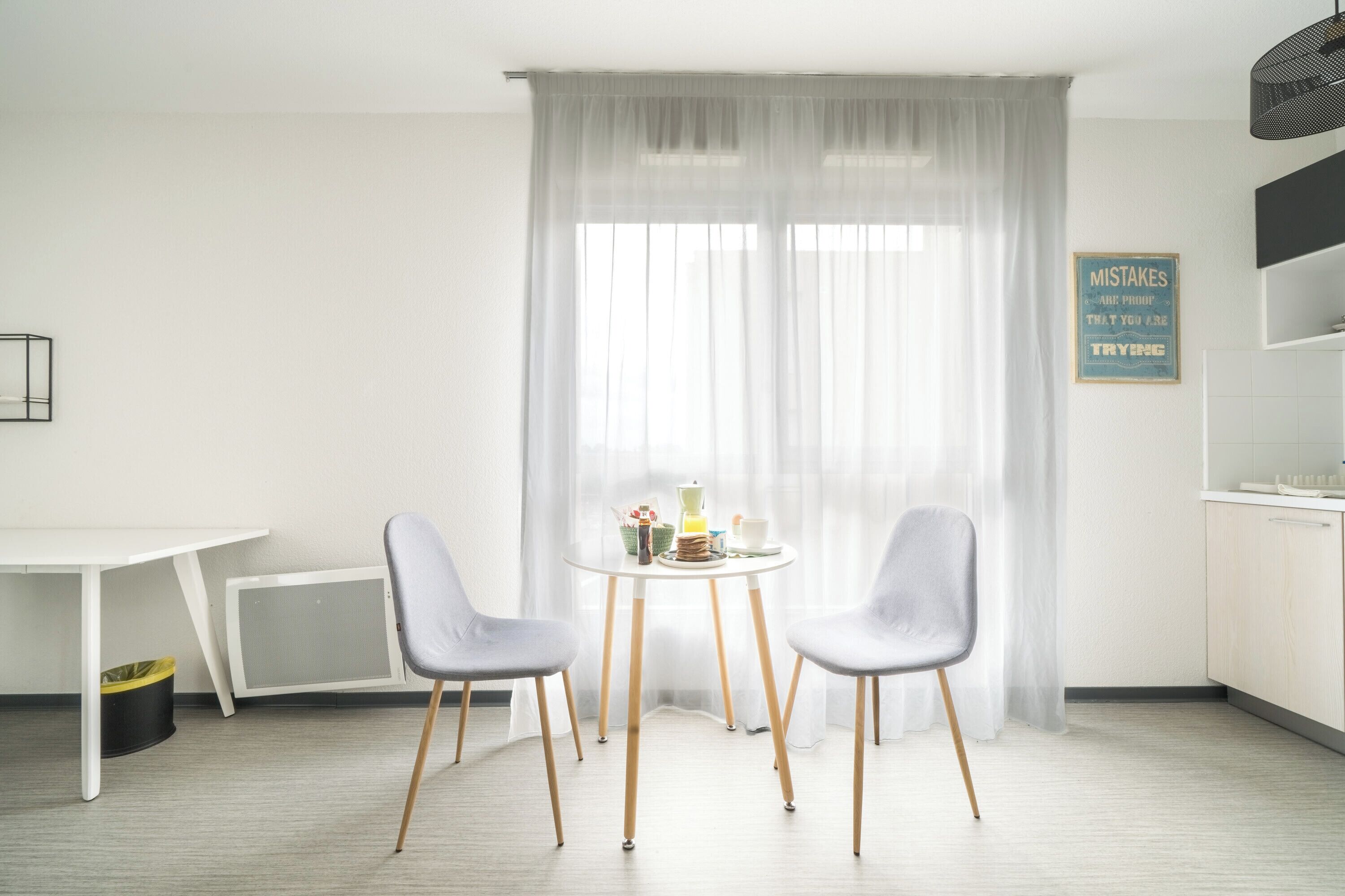 A modern room with a white table, two chairs, a window with sheer curtains, a wall-mounted sign, and a kitchen area.
