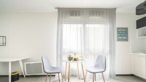 A modern room with a white table, two chairs, a window with sheer curtains, a wall-mounted sign, and a kitchen area. - A308 - Le Soléa - Wifi - Proche tram (Montpellier)