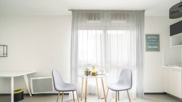 A modern room with a white table, two chairs, a window with sheer curtains, a wall-mounted sign, and a kitchen area.