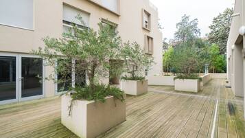 A courtyard with wooden flooring, planters with greenery, and a building with large windows and a door.