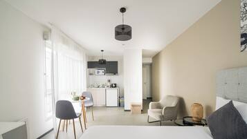 A modern hotel room with a bed, a desk, a chair, a small kitchen area, and a dining table.