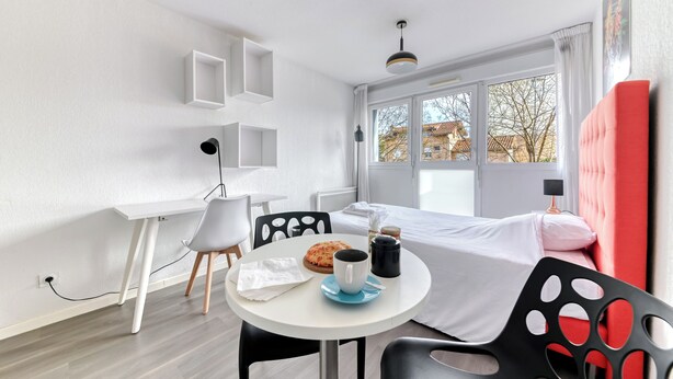 Une chambre d’hôtel moderne équipée d’un lit, d’un bureau, d’une chaise, d’une table avec une tasse et d’une fenêtre donnant sur des immeubles.