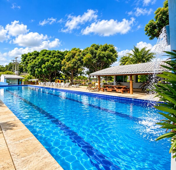 Una piscina con agua azul cristalina, rodeada de sillas de descanso, una zona de estar cubierta y abundante vegetación.