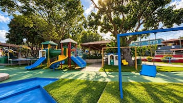 A playground with slides and swings, surrounded by trees and a clear sky.