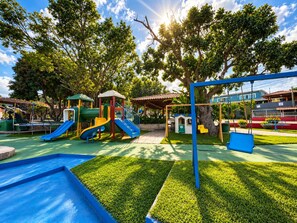 Un parque infantil con toboganes y columpios, rodeado de árboles y un cielo despejado.