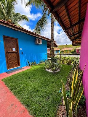A blue building with a wooden door, a small garden with grass and plants, and a palm tree. - Amoras Hotel Fazenda (Conceição do Almeida)