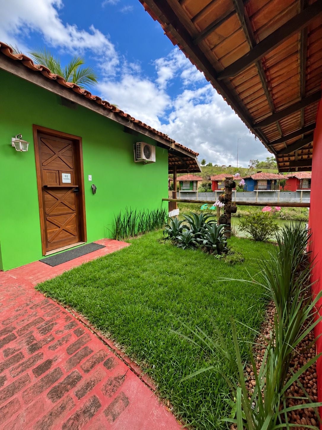 A green building with a wooden door, a red brick pathway, and a small garden area with plants and a decorative structure.