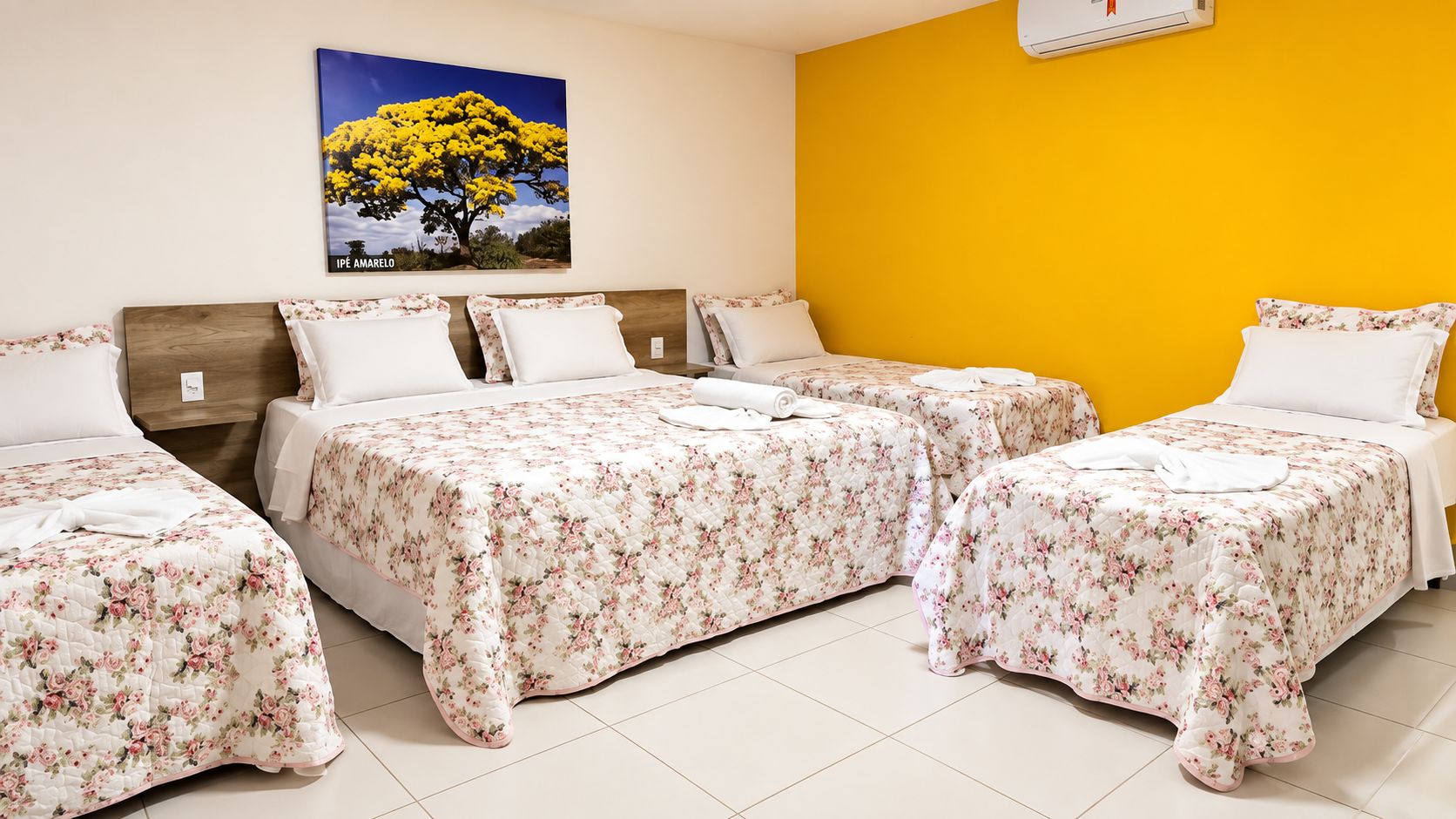 Une chambre d’hôtel avec trois lits, chacun recouvert d’une couvre-lit à motifs floraux, dotée d’une table de chevet, d’un climatiseur mural et d’un tableau représentant un arbre jaune.