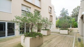 Een binnenplaats met houten vloer, plantenbakken met beplanting, en een gebouw met grote ramen en een deur.