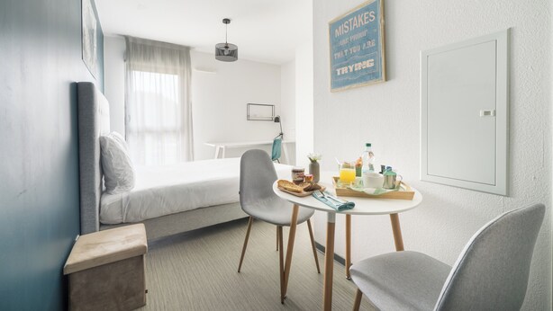 Moderni hotellihuone, jossa on sänky, aamiaistarvikkeilla katettu ruokapöytä, tuoli ja seinälle kiinnitetty kyltti.