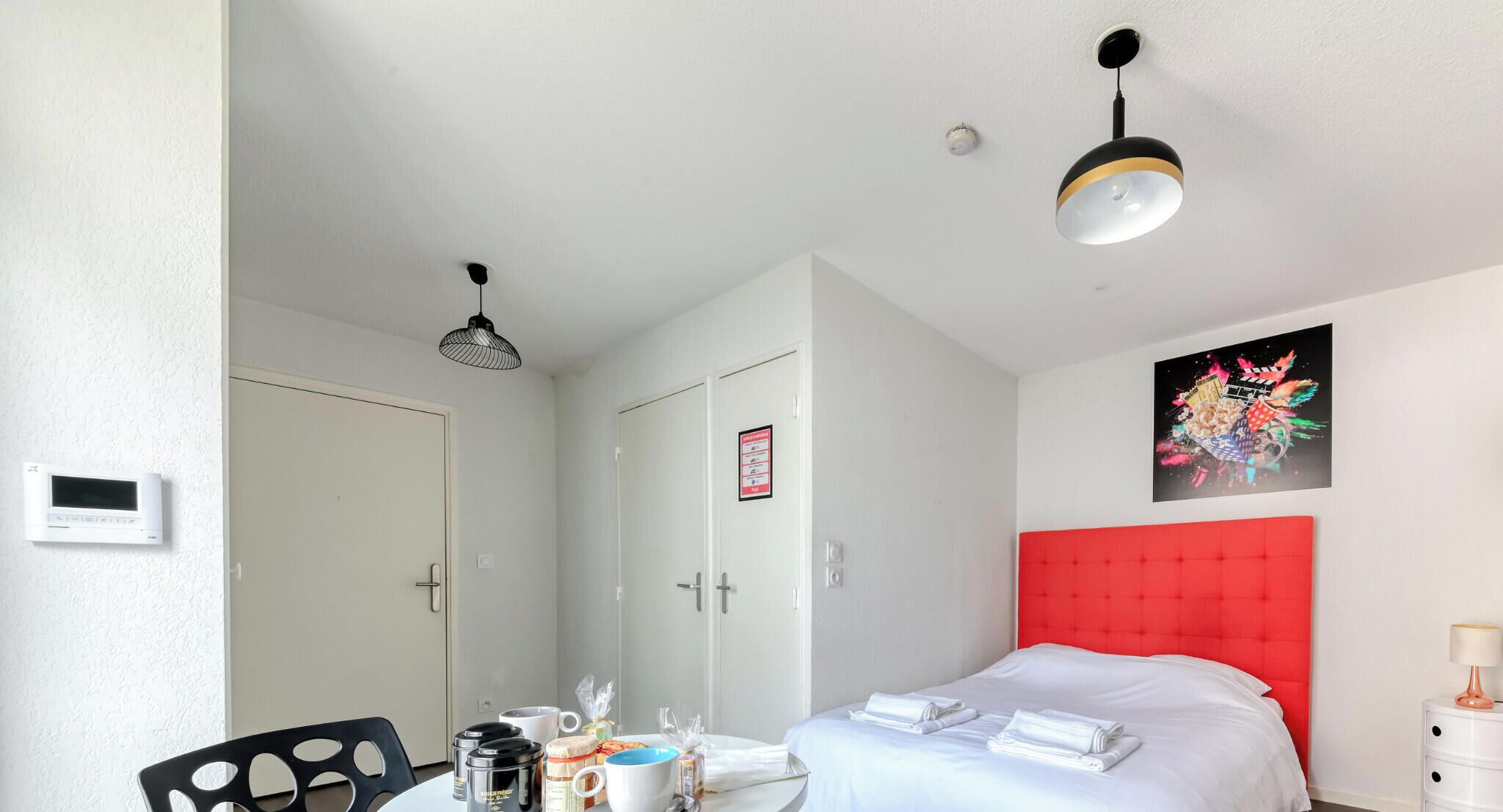 Ett hotellrum med en säng, ett matbord med frukosttillbehör, en röd sänggavel och ett konstverk som hänger på väggen.