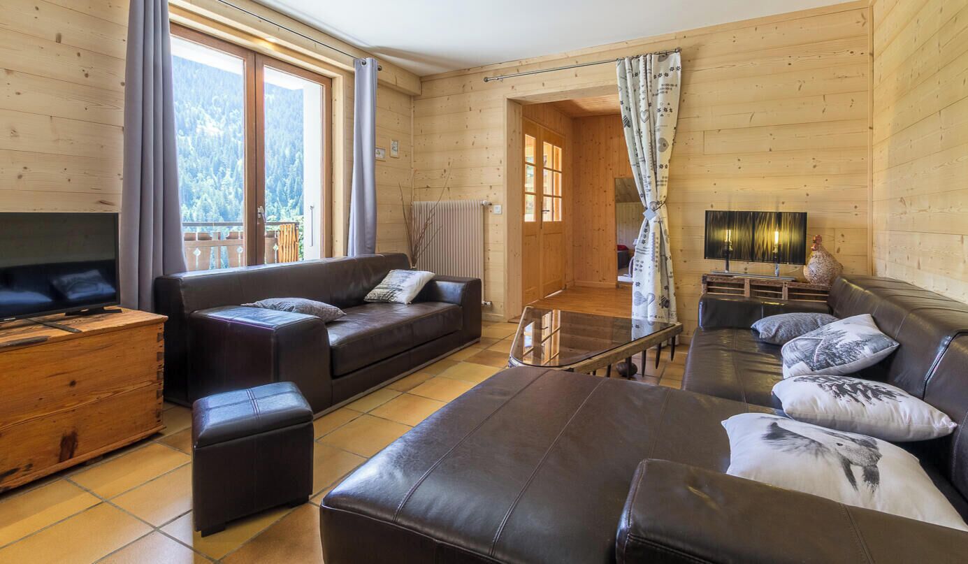 L’intérieur d’un chalet chaleureux, avec un canapé, un pouf et un téléviseur posé sur une commode en bois.