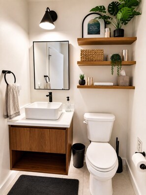 Een moderne badkamer met een witte wastafel, spiegel en toilet, houten planken met planten en toiletartikelen, en een zwarte prullenbak.