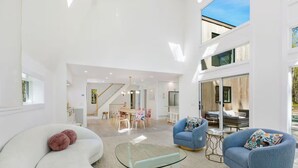 Un vaste espace de vie comprenant un canapé blanc incurvé, une table basse en verre et deux fauteuils bleus ornés de coussins à motifs.