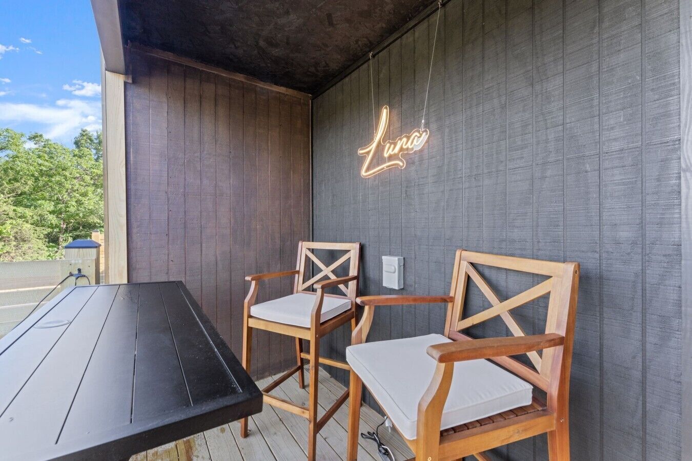 Un espacio de estar al aire libre con madera, dos sillas, una mesa, un letrero de neón y una pared gris.