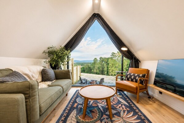 A cozy room with a sofa, a chair, a small table, a TV, and a large window offering a scenic view. - Luna A-Frame | King Suite, Fire Pit & Hot Tub (Berkeley Springs)