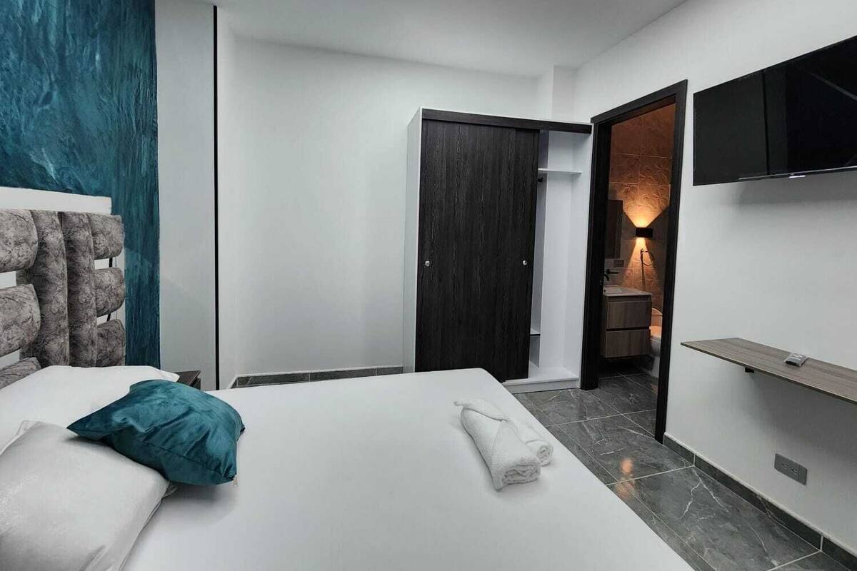 A modern hotel room with a bed, a TV, a bathroom, and a dark wooden door.