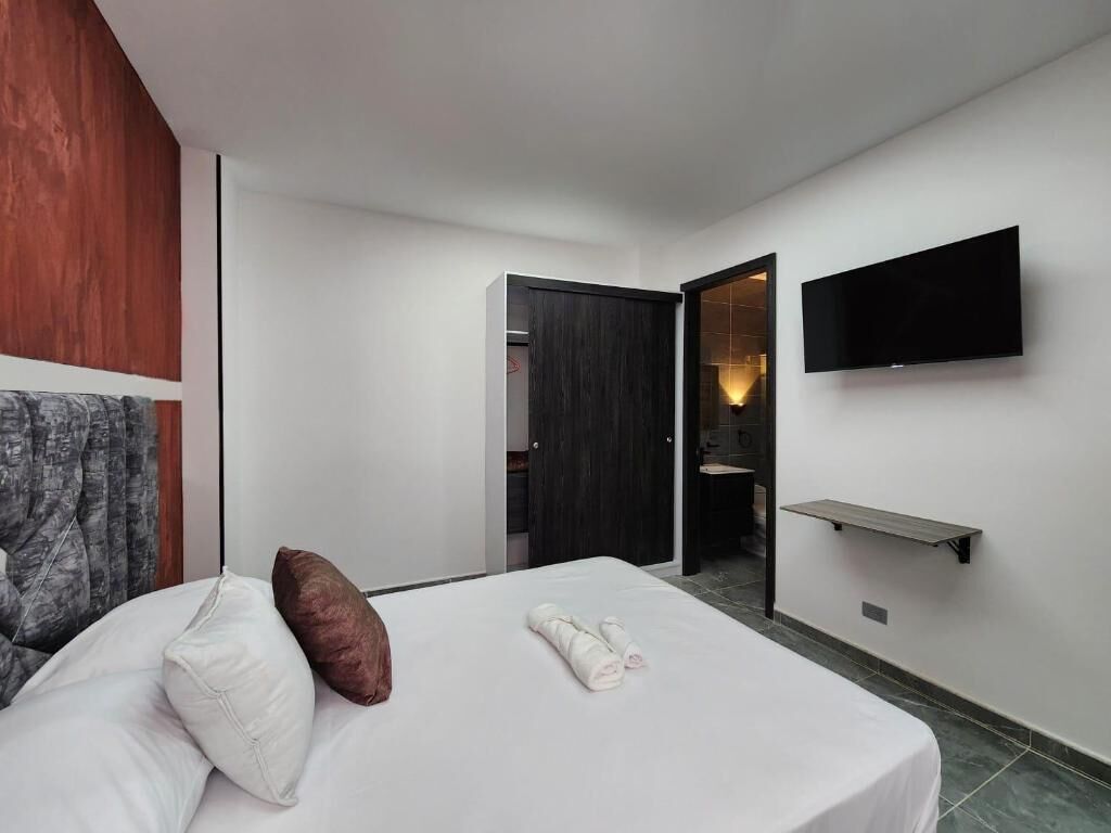 A hotel room with a bed, a TV mounted on the wall, a wooden panel on the left, and a door leading to a bathroom.