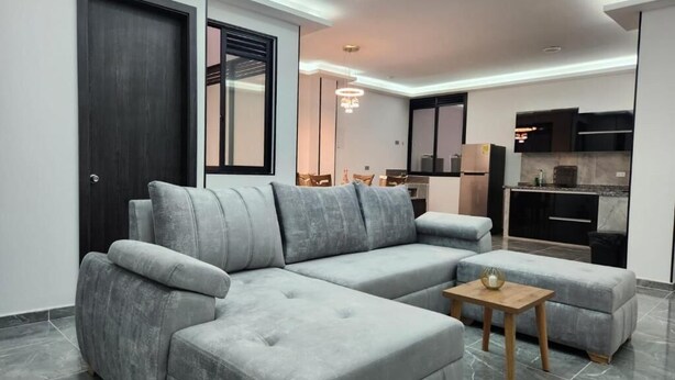 Una sala de estar moderna con un sofá seccional gris, una mesa de centro de madera y una cocina al fondo.
