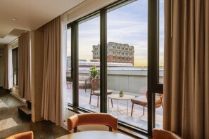 Ein Hotelzimmer mit großen Fenstern, Blick auf ein Gebäude, einem kleinen Balkon mit Sitzgelegenheit und einem Blumentopf.