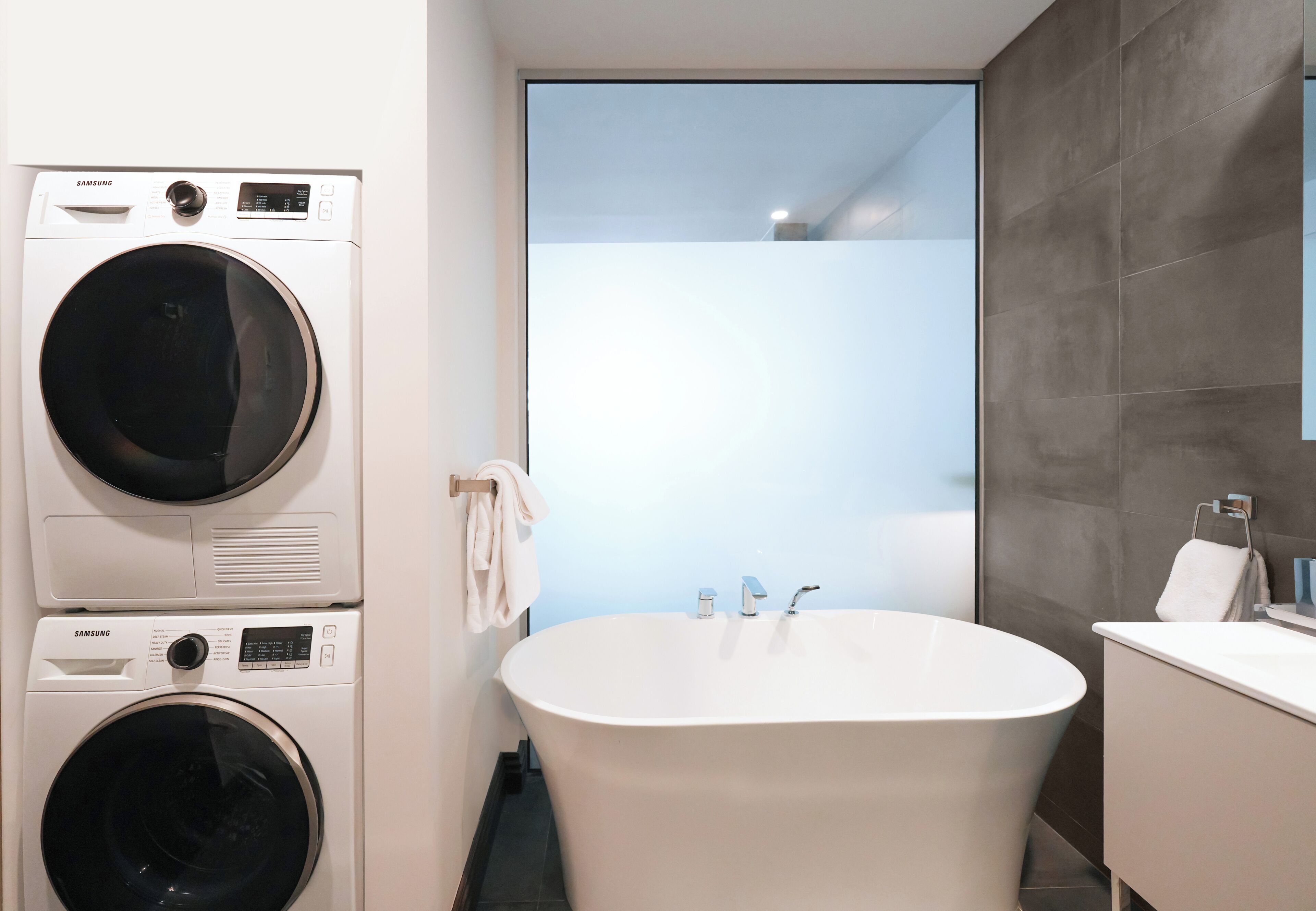 A modern bathroom with a built-in washer and dryer, a freestanding bathtub, and a large window.