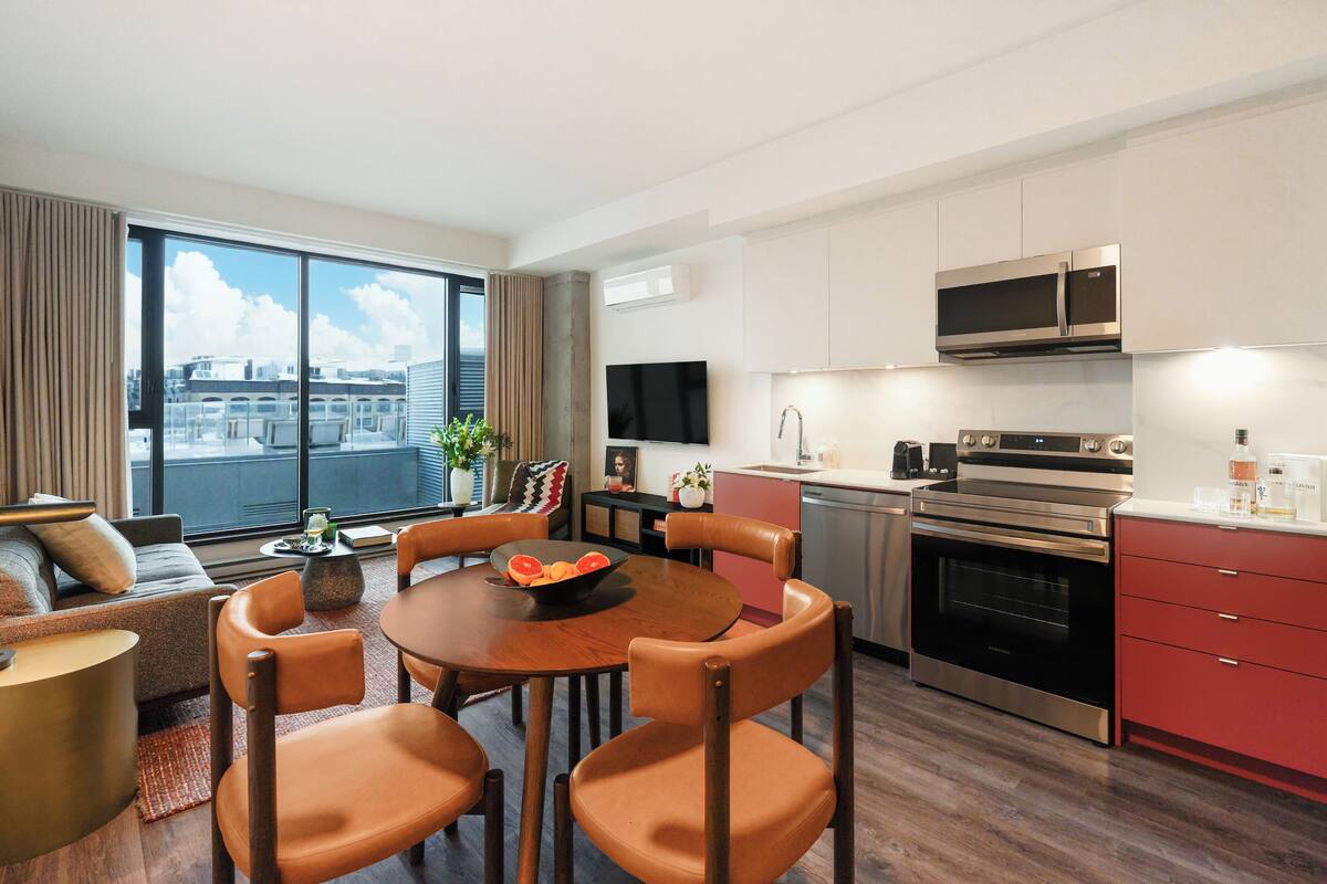 A modern hotel room with a dining area, kitchen, and large windows offering a city view.