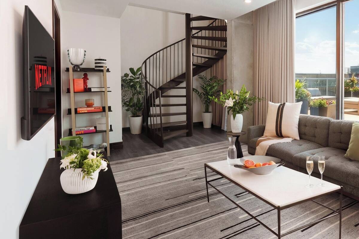 A modern living room with a flat-screen TV, a coffee table, a sofa, a staircase, and large windows.