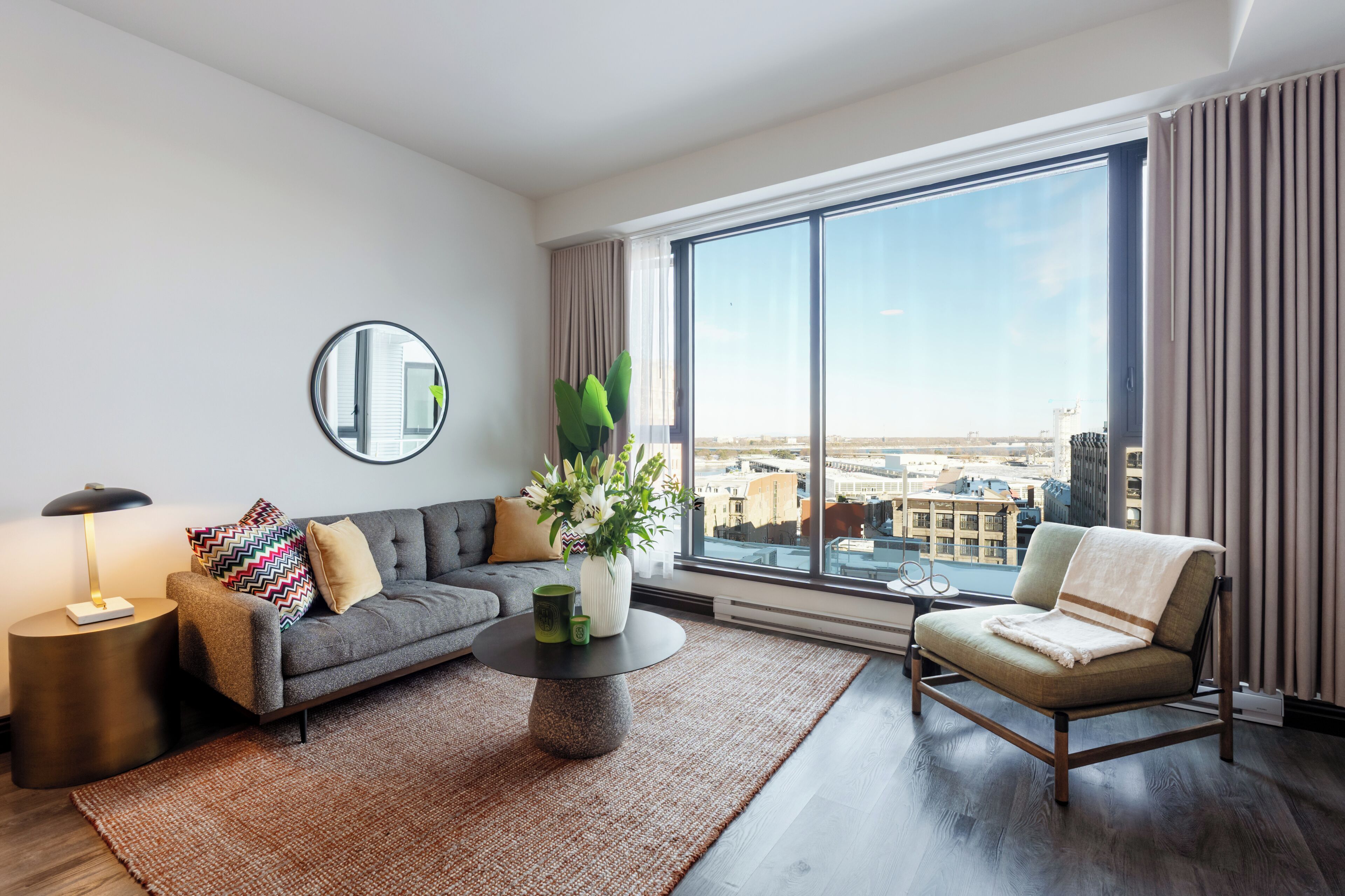 A modern living room with a sofa, armchair, round coffee table, and a large window with a city view.