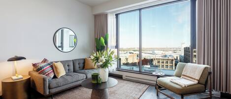 A modern living room with a sofa, armchair, round coffee table, and a large window with a city view.