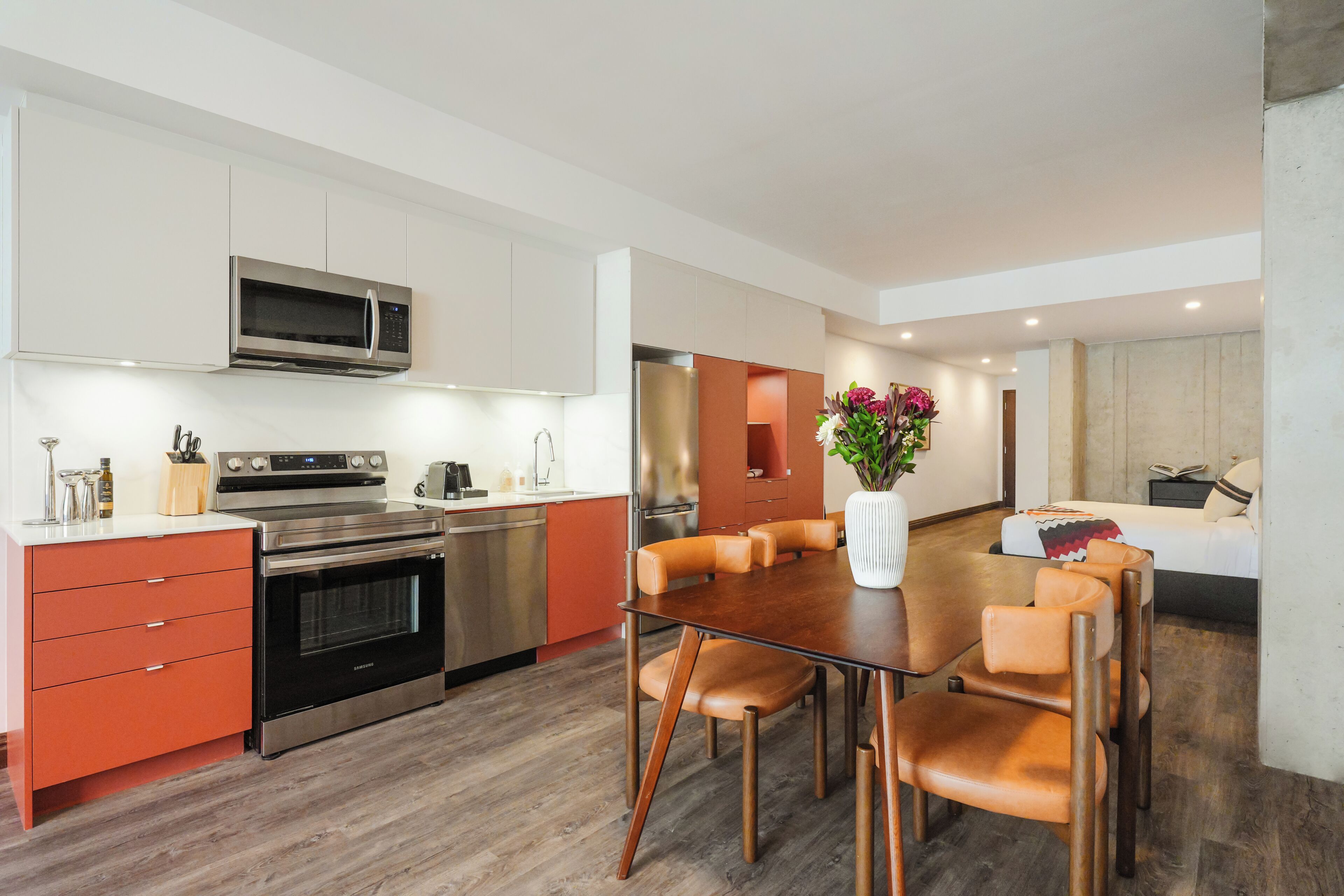 A modern kitchen with stainless steel appliances, a dining area with wooden chairs, and a living space with a sofa and a decorative vase.