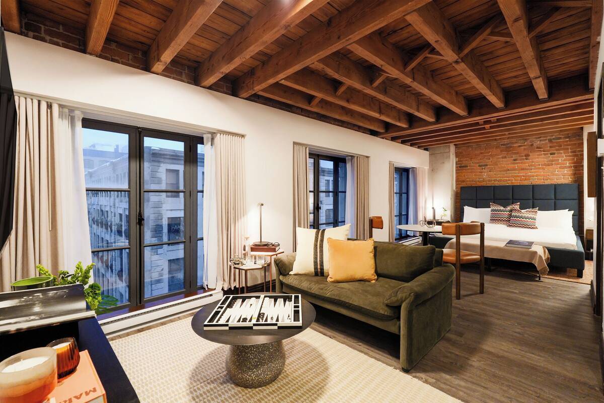 A hotel room with a wooden ceiling, a sofa, a bed, a small table, and a view through large windows.