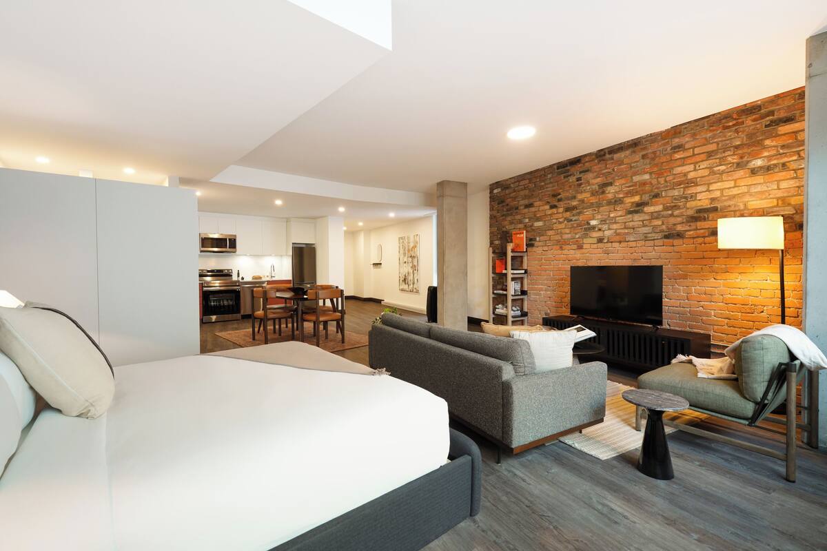 A modern hotel room with a bed, sofa, TV, fireplace, and a kitchen area in the background.