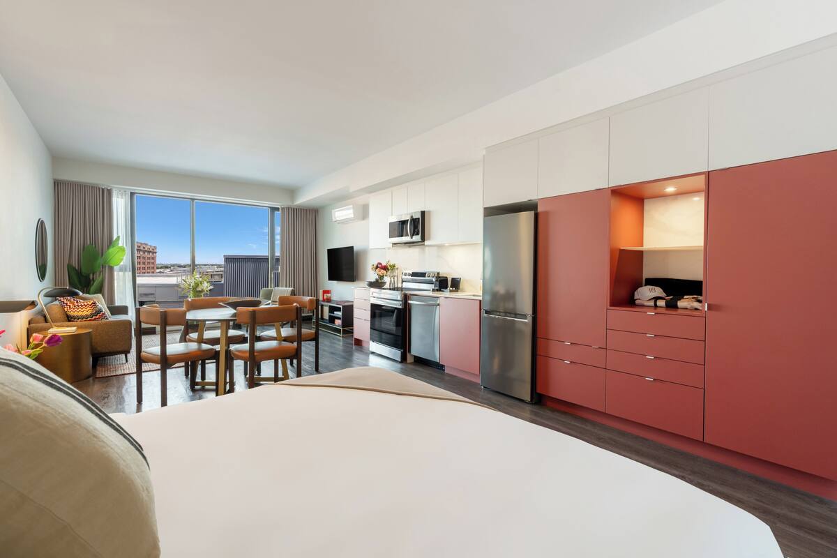 A modern hotel room with a dining area, kitchenette, and a large window offering a city view.