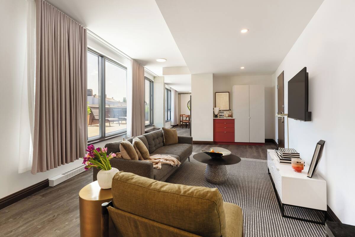 A modern hotel room with a living area, a sofa, a coffee table, a TV, and a view of the city.