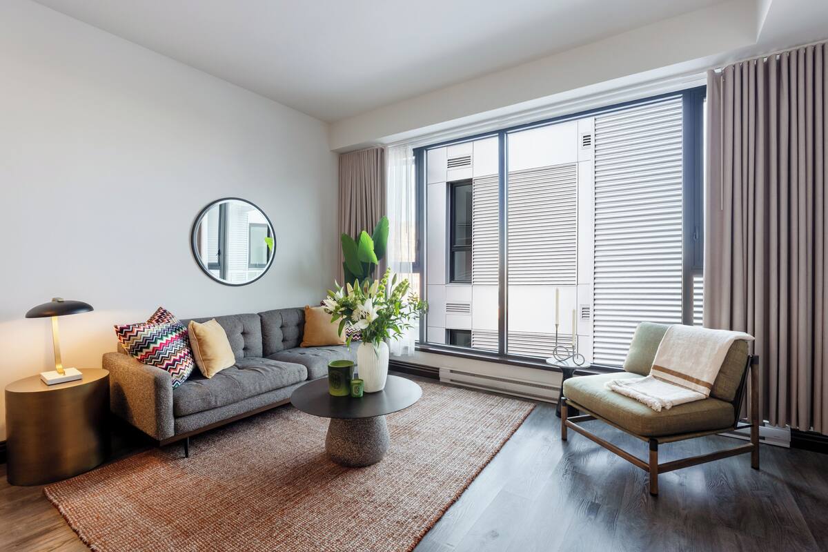 A modern living room with a grey sofa, a round coffee table, a side chair, a mirror, and a large window with blinds.