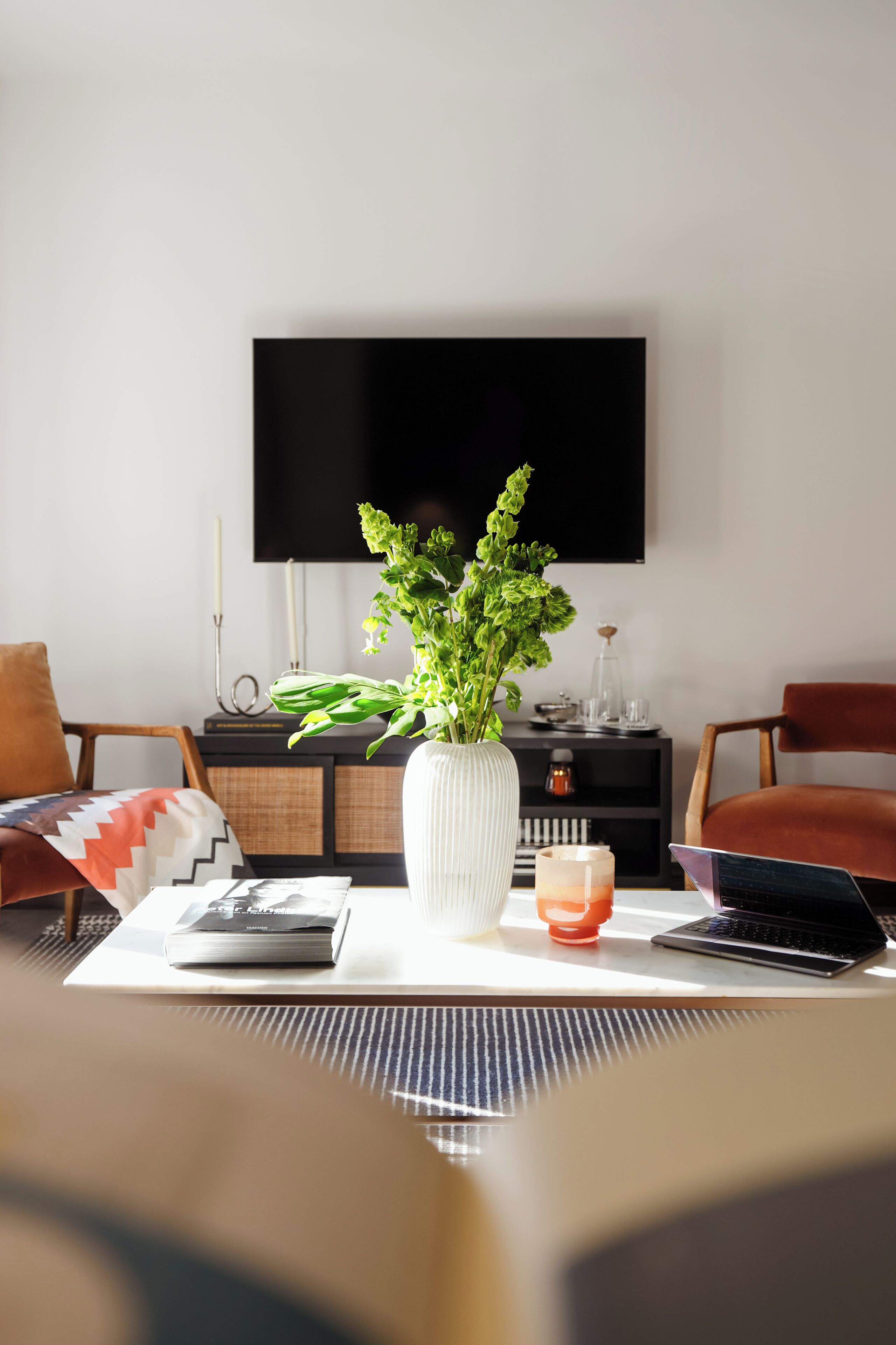 A modern living room with a flat-screen TV, a white vase with green plants, a laptop, and a coffee cup on a coffee table.