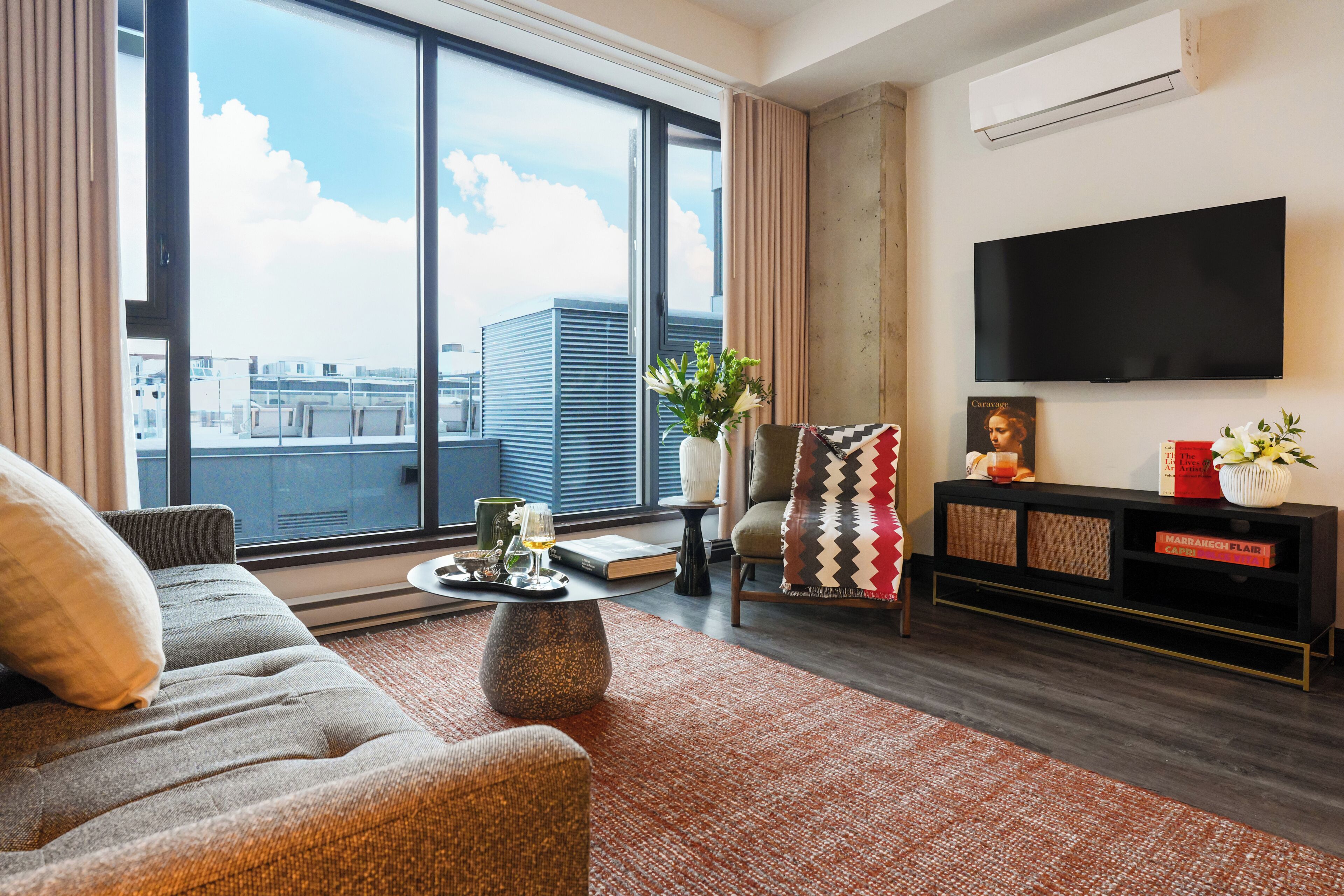 Una sala de estar moderna con un sofá, una mesa de centro, un televisor y un amplio ventanal con vista a la ciudad.
